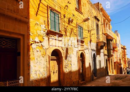 Alte verwitterte Straße in Malta mit goldenen Gebäuden, abblätternder Farbe, bogenförmigen Holztüren und grünen Fensterläden. Schatten, die von Sonnenlicht geworfen werden, erhöhen den Wert Stockfoto