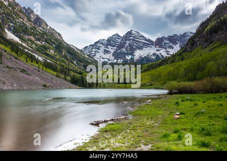 Majestätische Maroon Bells erheben sich über dem Maroon Lake, umgeben von üppigem Grün und schneebedeckten Gipfeln in Aspen, Colorado, im Frühling. Stockfoto