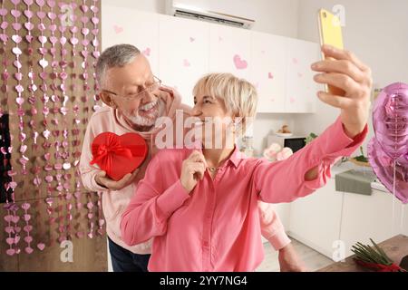 Glückliches reifes Paar mit Geschenkbox und Handy, das zu Hause Selfie in der Küche macht. Valentinstag-Feier Stockfoto