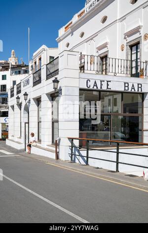 Eine Frau sitzt vor der Casino Bar im Hafen von Cadaqués in der Morgensonne in Katalonien, Spanien Stockfoto
