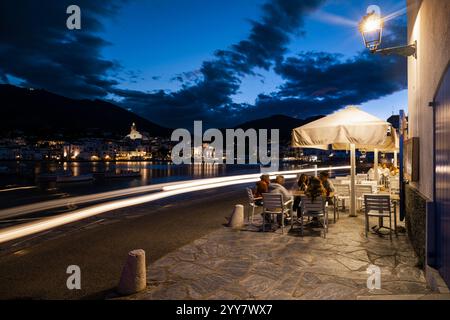 In Cafés sitzen Menschen und schlendern in der Abenddämmerung entlang der beleuchteten Uferpromenade im Hafen und in der Altstadt von Cadaqués, Katalonien, Spanien Stockfoto