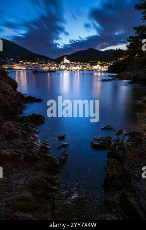 Blick von einer felsigen Bucht auf die beleuchtete Altstadt von Cadaqués in der Abenddämmerung, Katalonien, Spanien Stockfoto