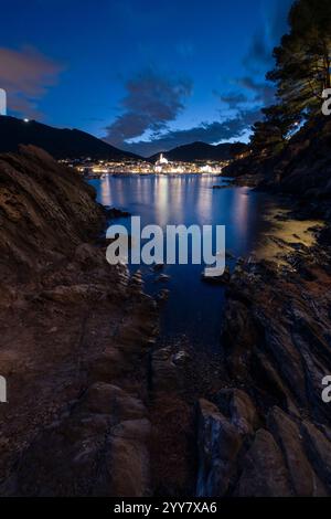 Blick von einer felsigen Bucht auf die beleuchtete Altstadt von Cadaqués in der Abenddämmerung, Katalonien, Spanien Stockfoto