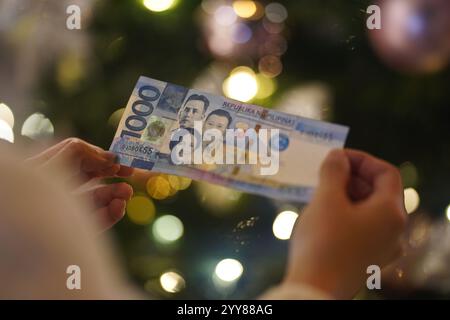 China. Dezember 2024. In dieser Abbildung hält eine Person philippinische Peso-Banknoten mit Weihnachtslichtern im Hintergrund. Quelle: SOPA Images Limited/Alamy Live News Stockfoto