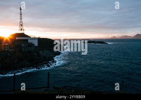 Ein malerischer Blick auf den Sonnenuntergang über einem nordischen Küstendorf, mit einem hohen Funkmast, der sich am Himmel befindet. Die felsige Küste und das ruhige Meer Stockfoto