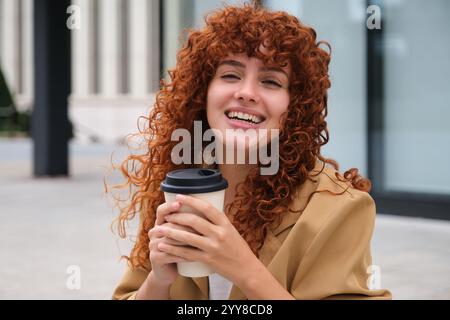 Glückliche rothaarige Geschäftsfrau, die eine Kaffeetasse hält und draußen lächelt Stockfoto