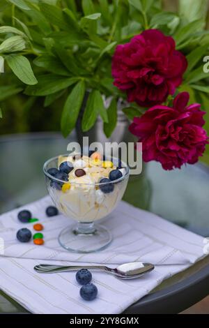 Köstliches Vanilleeis in einer Glasschale mit frischen Blaubeeren, bunten Süßigkeiten und Mandelscheiben auf einer weißen Serviette, mit leuchtenden roten Pfingstrosenblüten Stockfoto