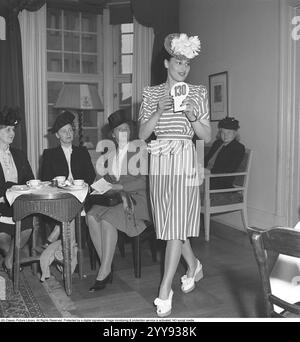 Modenschau 1946. Ein junges dunkelhaariges weibliches Model auf einer Modenschau. Sie trägt einen Anzug aus den 1940er Jahren mit einem passenden gestreiften Rock und einer hüftlangen Jacke. Sie läuft selbstbewusst in weißen High-Heels-Schuhen und einem schicken Hut und zeigt die Kleidung den Frauen, die sie beobachten. 1946 Kristoffersson Ref. T148-4 Stockfoto