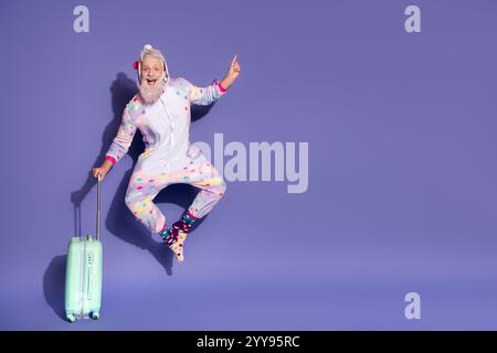 Fröhlicher Mann, der in Einhorn-Pyjamas springt, Koffer hält und auf violetten Hintergrund zeigt Stockfoto