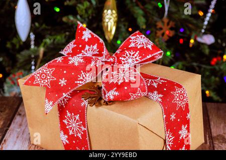 Wunderschön gewickeltes Geschenk mit rotem Schneeflockenband steht auf dem Tisch neben dem bunten Weihnachtsbaum während froher Weihnachten Stockfoto