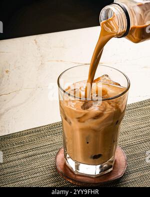Ein erfrischender Eiskaffee, der in ein mit Eiswürfeln gefülltes Glas gegossen wird, fängt den satten Wirbel und die geschmeidige Textur eines gekühlten, cremigen Getränks ein Stockfoto