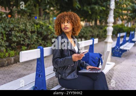 Lateinische junge Studentin mit Smartphone und Laptop, die auf der Parkbank sitzt Stockfoto