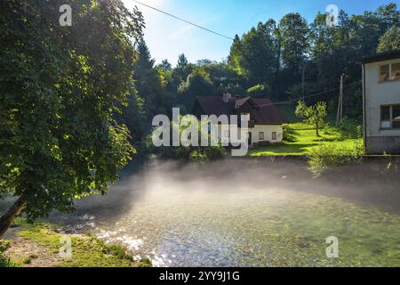 Das Sonnenlicht erhellt eine ruhige Szene, in der Morgennebel aus dem Fluss radovna in Bled, slowenien, aufsteigt, mit einem traditionellen Haus zwischen den Bäumen Stockfoto