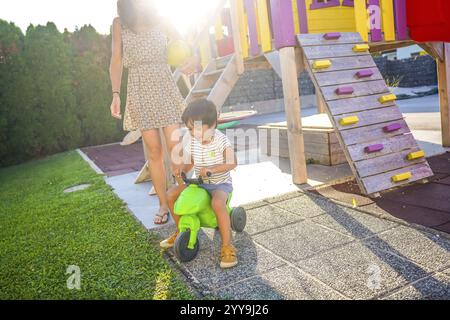 Der Junge fährt gern auf einem Spielplatz im Hinterhof auf einem Spielzeug-Motorrad, während seine Mutter an einem sonnigen Sommertag über ihn wacht Stockfoto