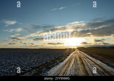 Eine ruhige Winterlandschaft mit einer unbefestigten Straße, die von schneebedeckten Feldern umgeben ist, unter einem glühenden Sonnenuntergang, der durch verstreute Wolken strömt, Stockfoto