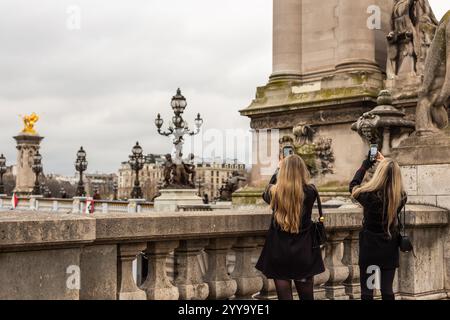 Zwei junge Frauen mit blonden Haaren fotografieren auf ihren I-Telefonen von der Brücke Pont Alexandre 111 über die seine. Eine wunderschöne Brücke in Paris Stockfoto