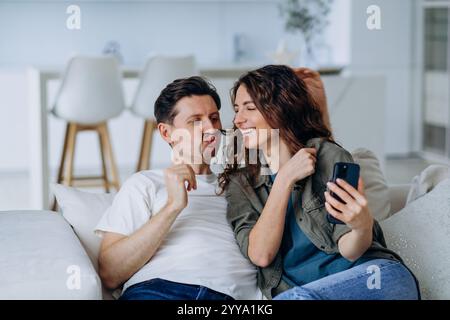 Ein junges Paar, das Spaß hat, ein Selfie auf dem Sofa zu Hause zu machen, der Mann macht ein lustiges Gesicht mit einem falschen Schnurrbart und die Frau lacht, sie benutzen ein Smartphone Stockfoto
