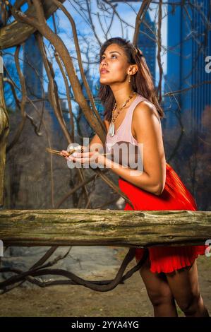 Eine Frau in einem leuchtend roten Kleid steht in einem Waldgebiet mit einem kleinen Eiernest und blickt nachdenklich nach oben. Urbane Wolkenkratzer erheben sich im Hintergrund Stockfoto