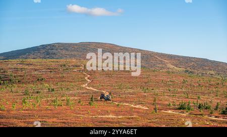 Zwei Geländefahrzeuge fahren auf einem kurvenreichen Bergpfad durch eine leuchtende rote herbstliche Tundra-Landschaft. Ein klarer blauer Himmel mit einer kleinen weißen Wolke unterstreicht die ruhige Landschaft Stockfoto