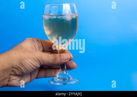 Nahaufnahme einer Hand mit einem transparenten Glas, gefüllt mit Weißwein, vor einem leuchtend blauen Hintergrund. Konzept der Feier, Alkohol, und r Stockfoto