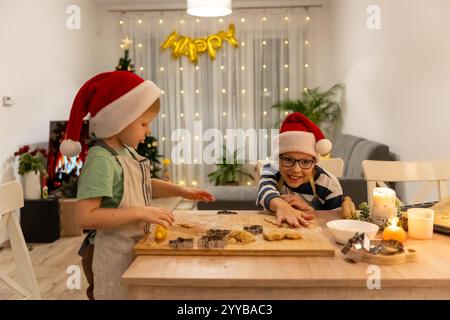 Zwei kleine Jungs in Weihnachtsmützen backen gemeinsam Weihnachtskekse zu Hause in gemütlicher festlicher Atmosphäre mit leuchtenden Lichtern und dekoriertem Weihnachtsfest Stockfoto