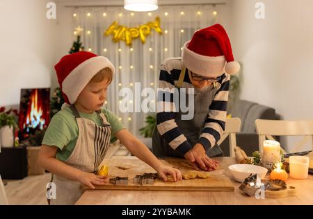 Zwei fröhliche Jungs mit Weihnachtsmütze, die Weihnachtskekse in der gemütlichen Küche machen, dekoriert mit festlichen Lichtern. Stockfoto