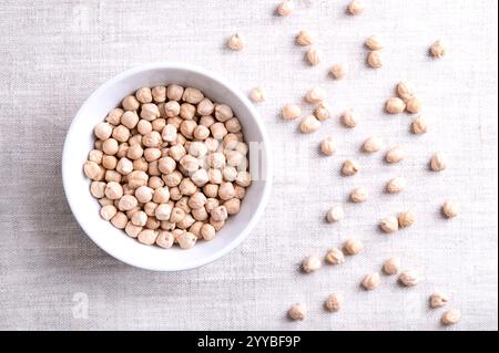 Getrocknete Kichererbsen in einer weißen Schüssel, auf Leinengewebe, von oben. Ganze und leicht gegerbte Kichererbsen, reich an Eiweißleguminosen und Früchten von Cicer arietinum. Stockfoto