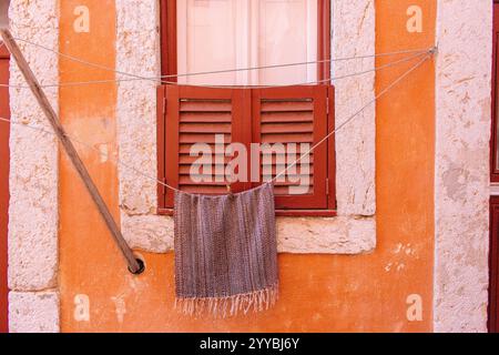 Trockentuch am Seil gegen orange verwitterte Wand. Geschlossenes Fenster mit Fensterläden an der alten Wand mit trocknender Bettwäsche. Gebäudefassade in Portugal. Stockfoto