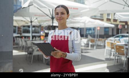 Junge hispanische Frau, die draußen auf der Terrasse des Cafés arbeitet und ein Tablett mit Schürze hält Stockfoto