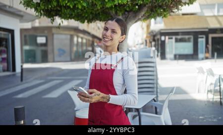 Eine junge Frau lächelt draußen auf einer Café-Terrasse und hält ein Telefon, trägt eine rote Schürze, steht vor gestapelten Stühlen auf einer Straße mit Bauwerk Stockfoto