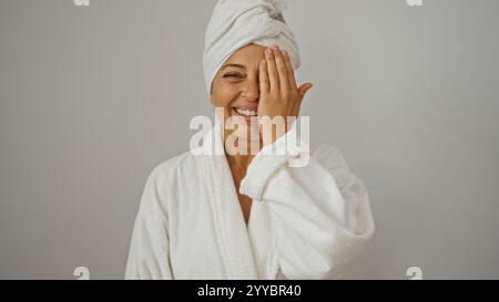 Eine Frau mittleren Alters mit einem Handtuch auf dem Kopf und im Bademantel bedeckt ein Auge mit der Hand, während sie vor weißem Hintergrund lächelt. Stockfoto
