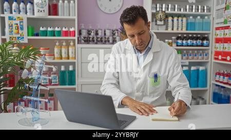 Mann, der drinnen in einer Apotheke arbeitet, während er einen Laptop benutzt und Notizen macht, umgeben von verschiedenen medizinischen Produkten und Hygieneartikeln in Ausstellungsregalen. Stockfoto