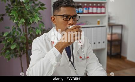 Doktor trinkt Getränk während einer Pause in einer Klinik, trägt Brille und einen weißen Mantel mit einem Stethoskop um den Hals in einem Innenarzt Stockfoto