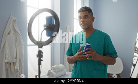 Junger hispanischer Mann in grünem Gewächs, der sich drinnen in einer Klinik mit Smartphone und Ringlicht filmt Stockfoto