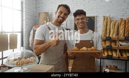 Zwei Bäcker in einer Bäckerei posieren zusammen mit frischen Croissants, tragen Schürzen, stehen drinnen, lächeln glücklich, zeigen Daumen hoch, Umrandung Stockfoto