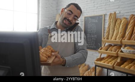 Junger Mann mit Schnurrbart, der in einer Bäckerei arbeitet, ein Tablett mit Gebäck hält, während er telefoniert, umgeben von verschiedenen Brotsorten in einem Innenraum Stockfoto