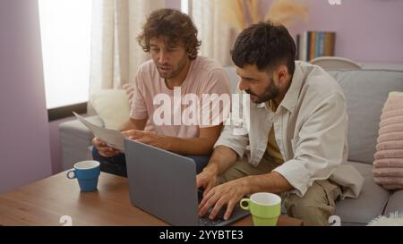 Zwei Männer sitzen in einem Wohnzimmer und arbeiten zusammen an einem Laptop mit Dokumenten und Kaffeetassen auf dem Tisch. So entsteht ein gemütliches und kooperatives Indoor-Set Stockfoto