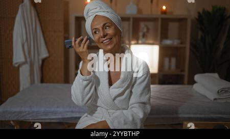 hispanische Frau mittleren Alters in einem Spa mit Bademantel und Handtuch, lächelnd während sie eine Sprachnachricht in einem ruhigen Wellnessraum hört. Stockfoto