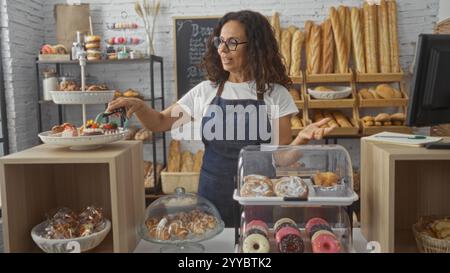 Frau, die hinter der Theke einer Bäckerei mit verschiedenen Backwaren und Brotsorten arbeitet, Schürze und Gläser trägt und Desserts mit t auswählt Stockfoto