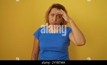 Eine hispanische Frau mittleren Alters in einem blauen Hemd mit lockigen Haaren steht isoliert vor einem gelben Hintergrund, sieht besorgt aus und berührt ihre Stirn. Stockfoto