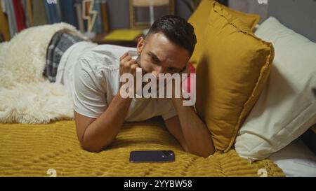 Ein junger hispanischer Mann mit Bart liegt auf einem Bett in einem Schlafzimmer, mit einem gelben Kissen und einem Handy neben ihm. Stockfoto