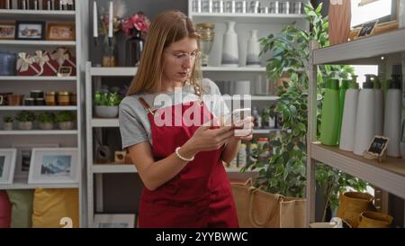 Frau, die ihr Telefon in einem Wohnkultur-Geschäft mit Regalen mit dekorativen Artikeln und Pflanzen um sie herum benutzt, die Produkte und eine lebendige Inneneinrichtung präsentieren Stockfoto