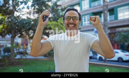 Mann, der fröhlich im Stadtpark mit einem Telefon mit lebhaftem Grün und Gebäuden im Hintergrund das Glück im Freien zeigt. Stockfoto