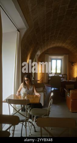 Junge Frau, die allein an einem Holztisch in einer schwach beleuchteten Hotelcafeteria mit rustikalen Bogendecken sitzt, Sonnenbrille und eine helle Bluse trägt, angenehm Stockfoto