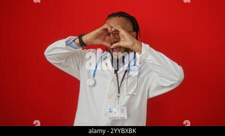 Ein Mann mit Zöpfen in einem weißen Mantel macht eine Herzgeste über einem isolierten roten Hintergrund, die einen fröhlichen und verspielten Ausdruck zeigt. Stockfoto