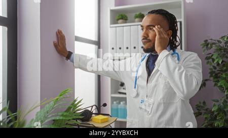 Doktor mit Dreadlocks in weißem Mantel lehnt sich an die Wand und berührt seinen Tempel in einem modernen Klinikraum, umgeben von Pflanzen und medizinischen Geräten Stockfoto