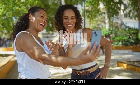 Zwei Frauen lächeln und winken während eines Videoanrufs in einem sonnigen Stadtpark und zeigen Freundschaft und Glück im Freien. Stockfoto