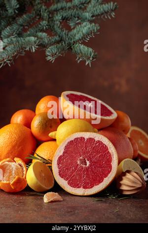 Zitrusfrüchte und Fichtenzweig auf braunem Hintergrund. Weihnachtliches Stillleben mit Orangen, Grapefruits, Zitronen und Mandarinen. Stockfoto