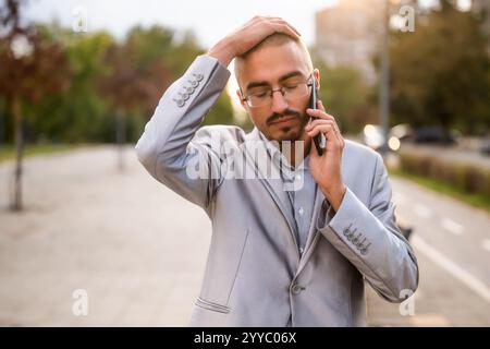 Porträt eines verzweifelten Geschäftsmannes, der am Telefon spricht. Junger Geschäftsmann, der auf der Straße steht und ein Smartphone benutzt. Stockfoto