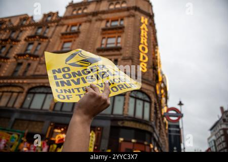 London, UK, 21. Dezember 2024. Ein Protestplakat befindet sich vor dem beleuchteten Harrods-Schild. Mitglieder der UVW union halten eine Demonstration vor dem weltberühmten Londoner Kaufhaus Harrods ab. Restaurantpersonal, Reinigungspersonal und Sicherheitsbeamte halten an einem der geschäftigsten Einkaufstage des Jahres eine Streikschlange vor dem Haupteingang. Die UVW-gewerkschaft fordert, dass ihren Mitgliedern ein versprochener Weihnachtsbonus, eine Erhöhung der Regellöhne und eine Verbesserung der allgemeinen Arbeitsbedingungen gezahlt wird. Quelle: James Willoughby/ALAMY Live News Stockfoto